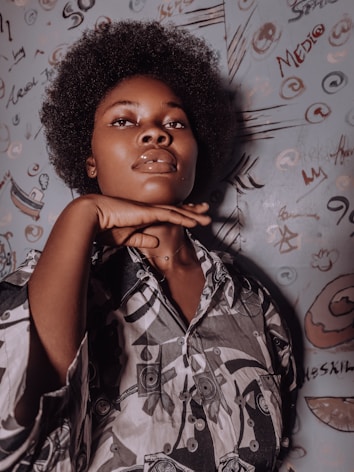 A person with an afro hairstyle poses confidently in front of a wall covered with abstract graffiti and doodles. The individual is wearing a patterned shirt and has a hand gracefully positioned under the chin.