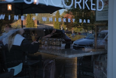 The image depicts the interior of a bar with a stone countertop and drinks set upon it. A reflection shows a person seated with a drink in hand, using a phone. The background reveals several parked cars and green trees, partially obscured by the glass reflection with bar signage.