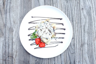 A white plate containing a dessert garnished with shredded coconut and chocolate drizzle. Fresh strawberries and a small sprig of mint add a pop of color. The plate rests on a rustic wooden surface.