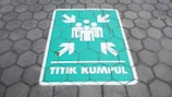 A painted sign on a hexagonal tile floor shows a green rectangle with white symbols. It contains human figures and arrows pointing inward, accompanied by the text 'TITIK KUMPUL'.
