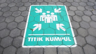 A painted sign on a hexagonal tile floor shows a green rectangle with white symbols. It contains human figures and arrows pointing inward, accompanied by the text 'TITIK KUMPUL'.