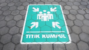 A painted sign on a hexagonal tile floor shows a green rectangle with white symbols. It contains human figures and arrows pointing inward, accompanied by the text 'TITIK KUMPUL'.