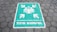A painted sign on a hexagonal tile floor shows a green rectangle with white symbols. It contains human figures and arrows pointing inward, accompanied by the text 'TITIK KUMPUL'.