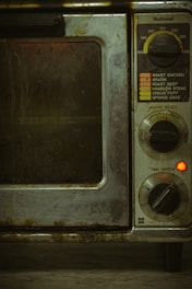 An old, well-used oven with dials and settings for different cooking temperatures and modes. The oven's metal exterior appears worn and dirty, likely from frequent use over time. There is a small red light indicating that part of the appliance is on.