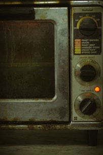 An old, well-used oven with dials and settings for different cooking temperatures and modes. The oven's metal exterior appears worn and dirty, likely from frequent use over time. There is a small red light indicating that part of the appliance is on.