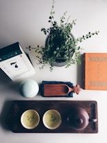A flat lay image featuring various objects on a light surface. In the center, there is a potted plant with green leaves. To the left, a box with text and a calendar-like design is positioned near a light-colored ceramic container. A wooden tray occupies the bottom area, holding a teapot and two teacups with painted designs. A wooden scoop rests on a dark cloth.