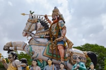 A detailed and colorful statue depicts a warrior seated on a richly adorned horse. The warrior is wearing ornate armor and a decorated headdress, holding a spear in one hand. Surrounding the main figure are smaller statues of various individuals, likely part of a procession or guard. The backdrop consists of a cloudy sky and patches of greenery.