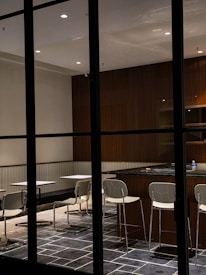 A modern, minimalistic dining area with tables and chairs arranged in a neat layout. The space features a dark tiled floor, wood-paneled walls, and overhead lighting that casts a warm glow. Chairs are positioned at each table, and a counter is visible with additional seating. A bottled water and a small object are placed on the counter.