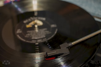 A close-up of a vinyl record on a turntable with the tonearm positioned over the record. The record has a reflective surface with visible grooves and faint text on the label.