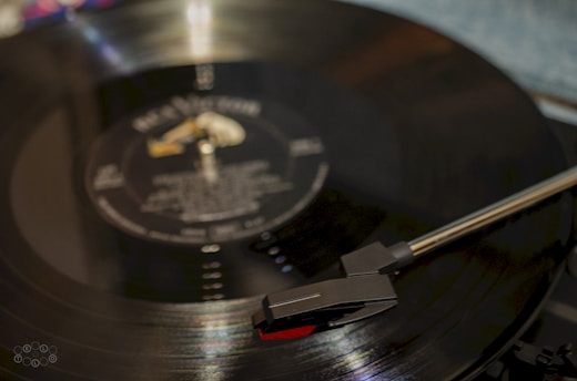 A close-up of a vinyl record on a turntable with the tonearm positioned over the record. The record has a reflective surface with visible grooves and faint text on the label.