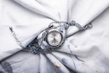 A silver wristwatch with a delicate chain is placed on a softly textured white fabric. The watch features a round face and the chain is adorned with small blue gemstones, adding a touch of elegance. The fabric is slightly wrinkled, creating a backdrop that enhances the watch's intricate details.