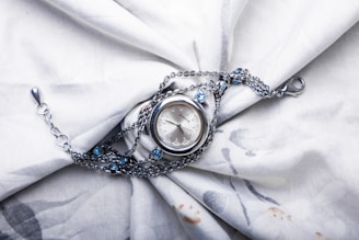A silver wristwatch with a delicate chain is placed on a softly textured white fabric. The watch features a round face and the chain is adorned with small blue gemstones, adding a touch of elegance. The fabric is slightly wrinkled, creating a backdrop that enhances the watch's intricate details.