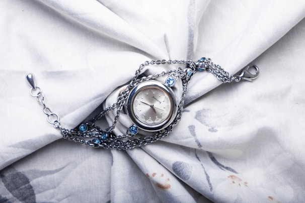 A silver wristwatch with a delicate chain is placed on a softly textured white fabric. The watch features a round face and the chain is adorned with small blue gemstones, adding a touch of elegance. The fabric is slightly wrinkled, creating a backdrop that enhances the watch's intricate details.