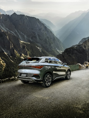 A modern SUV parked on a mountain road with rugged, towering cliffs and a dramatic valley view. The vehicle's sleek and polished design contrasts with the natural, rugged terrain. Sunlight filters through the mist, casting ethereal rays across the valley.