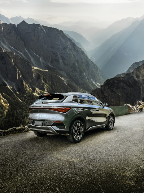 A modern SUV parked on a mountain road with rugged, towering cliffs and a dramatic valley view. The vehicle's sleek and polished design contrasts with the natural, rugged terrain. Sunlight filters through the mist, casting ethereal rays across the valley.