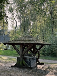 An old wooden well stands in a serene forest setting, surrounded by tall trees with lush green leaves. The well has a rustic wooden roof and a large wheel, situated on a cobblestone path. Sunlight filters through the foliage, casting dappled shadows on the ground.