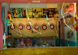 A booth featuring archery targets aligned in a row against a backdrop. Several toy bows and arrows are on display, hanging above the targets. A framed poster showing people engaging in archery is above the targets. Below the targets, there are various stuffed toys and other miscellaneous items neatly arranged. A person is seated on the left side, possibly the booth operator. On the right side, a fan and more toys are visible.