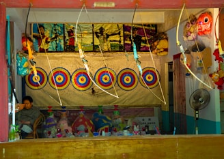 A booth featuring archery targets aligned in a row against a backdrop. Several toy bows and arrows are on display, hanging above the targets. A framed poster showing people engaging in archery is above the targets. Below the targets, there are various stuffed toys and other miscellaneous items neatly arranged. A person is seated on the left side, possibly the booth operator. On the right side, a fan and more toys are visible.
