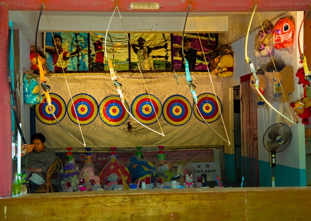 A booth featuring archery targets aligned in a row against a backdrop. Several toy bows and arrows are on display, hanging above the targets. A framed poster showing people engaging in archery is above the targets. Below the targets, there are various stuffed toys and other miscellaneous items neatly arranged. A person is seated on the left side, possibly the booth operator. On the right side, a fan and more toys are visible.