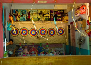 A booth featuring archery targets aligned in a row against a backdrop. Several toy bows and arrows are on display, hanging above the targets. A framed poster showing people engaging in archery is above the targets. Below the targets, there are various stuffed toys and other miscellaneous items neatly arranged. A person is seated on the left side, possibly the booth operator. On the right side, a fan and more toys are visible.