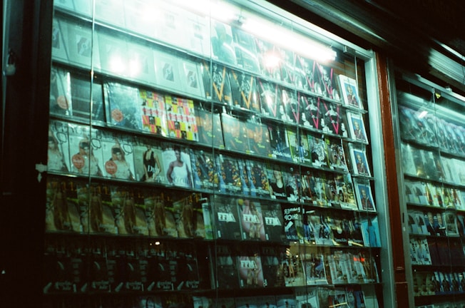 A storefront window display featuring an array of magazines. The magazines are arranged neatly in rows and columns, showcasing various covers with images and text. The lighting is slightly dim, reflecting off the glass window, and creates an urban, late-night atmosphere.