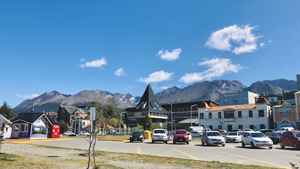 A small town is nestled at the base of a mountain range under a clear blue sky. Several cars are parked along the street, and a few trees and buildings are visible. The architecture includes a mix of modern and quaint structures, some with pointed roofs. A few clouds float gently in the sky.