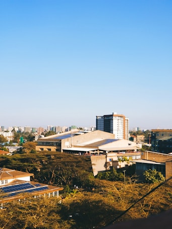 A cityscape featuring a mix of residential and commercial buildings. The foreground includes several buildings with flat roofs, one of which has solar panels. Trees dominate the lower part of the scene, while taller buildings are scattered in the background against a clear blue sky.