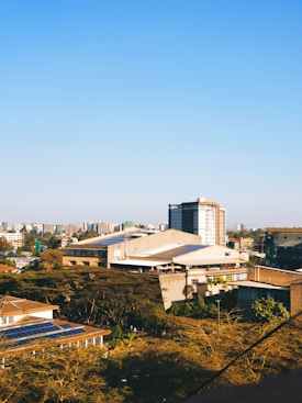 A cityscape featuring a mix of residential and commercial buildings. The foreground includes several buildings with flat roofs, one of which has solar panels. Trees dominate the lower part of the scene, while taller buildings are scattered in the background against a clear blue sky.