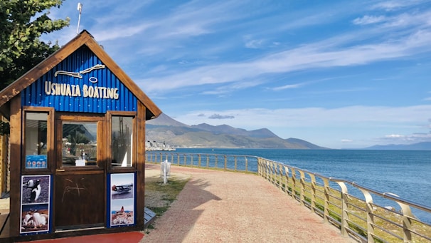 A wooden booth labeled 'Ushuaia Boating' stands near a waterfront. The structure features a blue, corrugated roof and is adorned with photographs of marine activities and wildlife. A rail-guarded walkway curves alongside the ocean, leading the eye toward distant mountains under a clear sky.