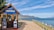 A wooden booth labeled 'Ushuaia Boating' stands near a waterfront. The structure features a blue, corrugated roof and is adorned with photographs of marine activities and wildlife. A rail-guarded walkway curves alongside the ocean, leading the eye toward distant mountains under a clear sky.