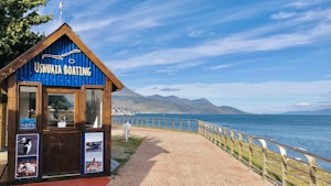 A wooden booth labeled 'Ushuaia Boating' stands near a waterfront. The structure features a blue, corrugated roof and is adorned with photographs of marine activities and wildlife. A rail-guarded walkway curves alongside the ocean, leading the eye toward distant mountains under a clear sky.