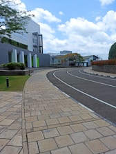A modern architectural setting with a curved pathway made of light-colored tiles. On the left, a building with geometric designs and greenery is visible, while lush trees and bushes enhance the landscape. The sky is partly cloudy, adding brightness to the scene.