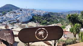 A picturesque hilltop village with white buildings and a lush green landscape. The view includes a large sign with a symbol and the word 'restaurant' partially visible. In the background, the village is surrounded by mountains and the sea is visible in the distance under a clear blue sky.