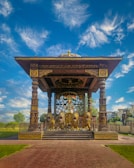 An ornate pavilion with intricate design elements features a central statue of a warrior riding a chariot pulled by four lions. The structure is supported by decorative columns and is richly detailed with patterns in gold and dark colors. Surrounding the pavilion are well-maintained lawns and trees under a vibrant blue sky with scattered clouds.