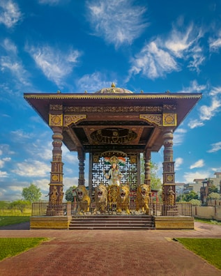 An ornate pavilion with intricate design elements features a central statue of a warrior riding a chariot pulled by four lions. The structure is supported by decorative columns and is richly detailed with patterns in gold and dark colors. Surrounding the pavilion are well-maintained lawns and trees under a vibrant blue sky with scattered clouds.