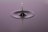 A droplet of water captured in mid-air as it descends towards a ripple created on a calm surface. The droplet creates a sharp, reflective peak that emerges from the smooth surface, with concentric circles emanating from the point of impact. The background has a soft gradient, enhancing the focus on the droplet.