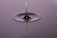 A droplet of water captured in mid-air as it descends towards a ripple created on a calm surface. The droplet creates a sharp, reflective peak that emerges from the smooth surface, with concentric circles emanating from the point of impact. The background has a soft gradient, enhancing the focus on the droplet.