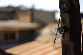 An old light bulb is attached to a weathered wooden post outside, with its base connected to a rustic power outlet. The background is blurred, indicating a rooftop and some structures in an urban setting.