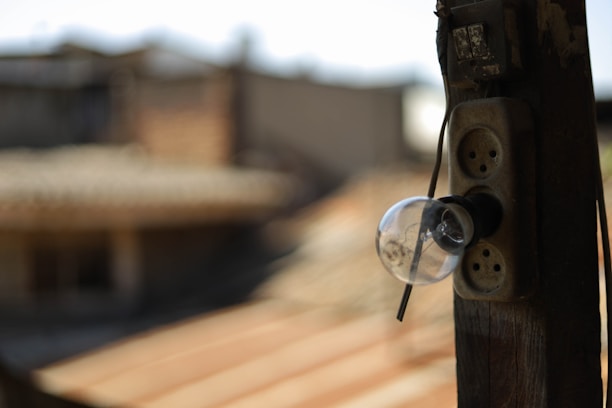 An old light bulb is attached to a weathered wooden post outside, with its base connected to a rustic power outlet. The background is blurred, indicating a rooftop and some structures in an urban setting.