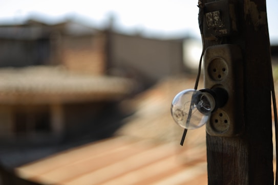 An old light bulb is attached to a weathered wooden post outside, with its base connected to a rustic power outlet. The background is blurred, indicating a rooftop and some structures in an urban setting.