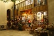 A cozy, warmly lit shop with a rustic wooden exterior, showcasing a variety of traditional handicrafts and decorative items. Colorful textiles, baskets, and dolls are displayed outside, inviting visitors to explore inside where more goods are visible through the glass windows. The scene is serene with a hint of nostalgia.