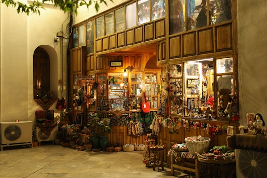 A cozy, warmly lit shop with a rustic wooden exterior, showcasing a variety of traditional handicrafts and decorative items. Colorful textiles, baskets, and dolls are displayed outside, inviting visitors to explore inside where more goods are visible through the glass windows. The scene is serene with a hint of nostalgia.