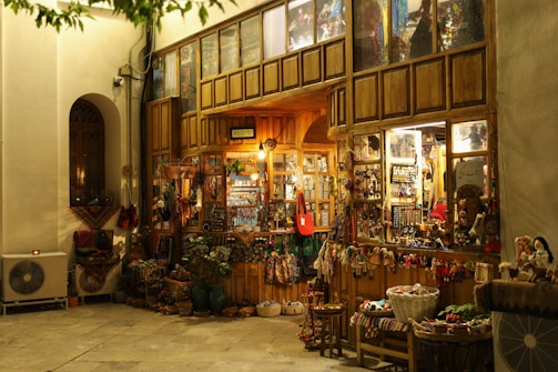 A cozy, warmly lit shop with a rustic wooden exterior, showcasing a variety of traditional handicrafts and decorative items. Colorful textiles, baskets, and dolls are displayed outside, inviting visitors to explore inside where more goods are visible through the glass windows. The scene is serene with a hint of nostalgia.
