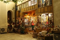 A cozy, warmly lit shop with a rustic wooden exterior, showcasing a variety of traditional handicrafts and decorative items. Colorful textiles, baskets, and dolls are displayed outside, inviting visitors to explore inside where more goods are visible through the glass windows. The scene is serene with a hint of nostalgia.
