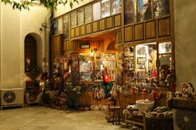 A cozy, warmly lit shop with a rustic wooden exterior, showcasing a variety of traditional handicrafts and decorative items. Colorful textiles, baskets, and dolls are displayed outside, inviting visitors to explore inside where more goods are visible through the glass windows. The scene is serene with a hint of nostalgia.
