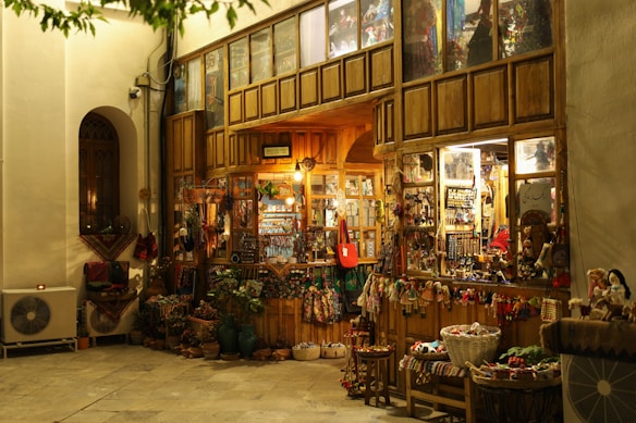 A cozy, warmly lit shop with a rustic wooden exterior, showcasing a variety of traditional handicrafts and decorative items. Colorful textiles, baskets, and dolls are displayed outside, inviting visitors to explore inside where more goods are visible through the glass windows. The scene is serene with a hint of nostalgia.