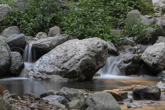 A serene natural scene featuring a small waterfall flowing over large, smooth rocks into a calm stream. The background is filled with lush, green foliage, and the water appears silky due to a long exposure effect.