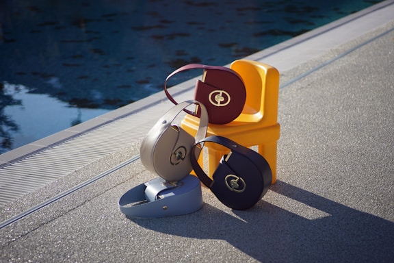 Several stylish handbags are arranged on a small yellow chair beside a pool. The bags feature a circular design and metallic logo. The setting is outdoors on textured pavement, with the water reflecting patterns from the sky.