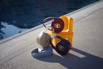 Several stylish handbags are arranged on a small yellow chair beside a pool. The bags feature a circular design and metallic logo. The setting is outdoors on textured pavement, with the water reflecting patterns from the sky.