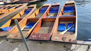 Wooden boats with blue cushions are aligned on a dock, chained and ready for use on the water. Each boat has a unique identification number and a paddle rests across one of them.